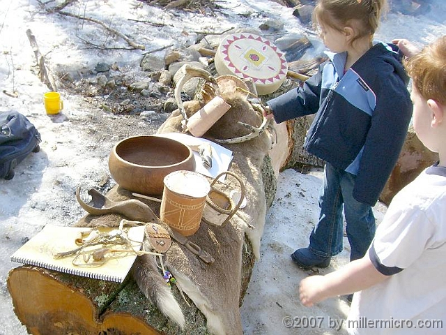 070303WampanoagSpoonEtAl1 The exhibit log holds a furry deer hide, a wooden bowl and birch-bark baskets, a deerskin drum, and more for kids to touch and feel. In front on the notebook, a toy made of a deer's leg bone and some of its (hollow) toe bones, strung together with a deer-hide thong. The game is to hold the leg bone sharp end up, and use it to swing the toe bones into the air. It's not easy to skewer one of them on the leg bone!  Just behind this toy, is an strange-looking piece of dark wood which is...